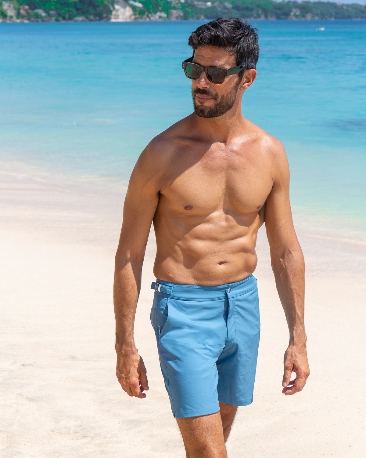 Man styled for the beach wearing RWENZORI Mykonos Blue Performance Series swim shorts with blue ocean water and white sand background.