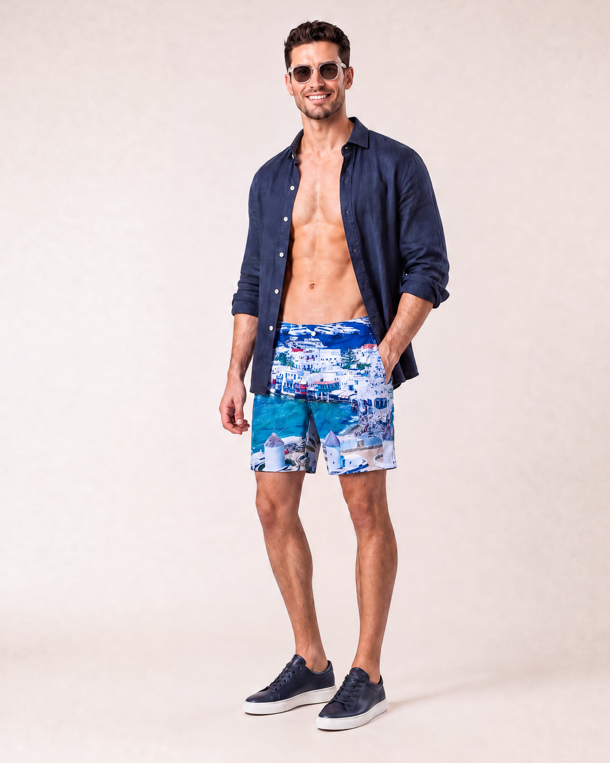 Man wearing RWENZORI Mykonos photo print swim shorts with an open navy linen resort shirt, three-quarter view.