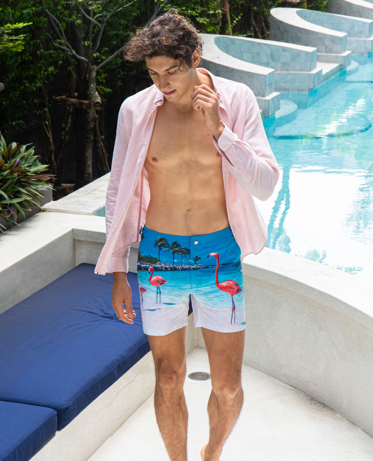 Man wearing Rwenzori flamingo swim shorts poolside lounge in linen shirt.