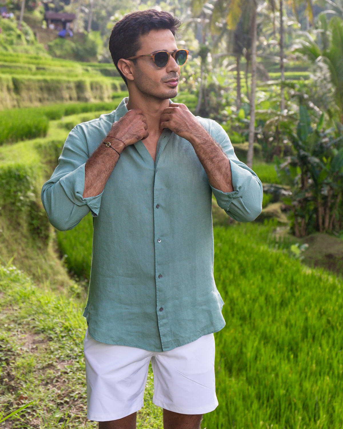 A man wearing RWENZORI sage green linen button-down shirt and white shorts stands in front of lush green terraced rice fields, adjusting his collar.