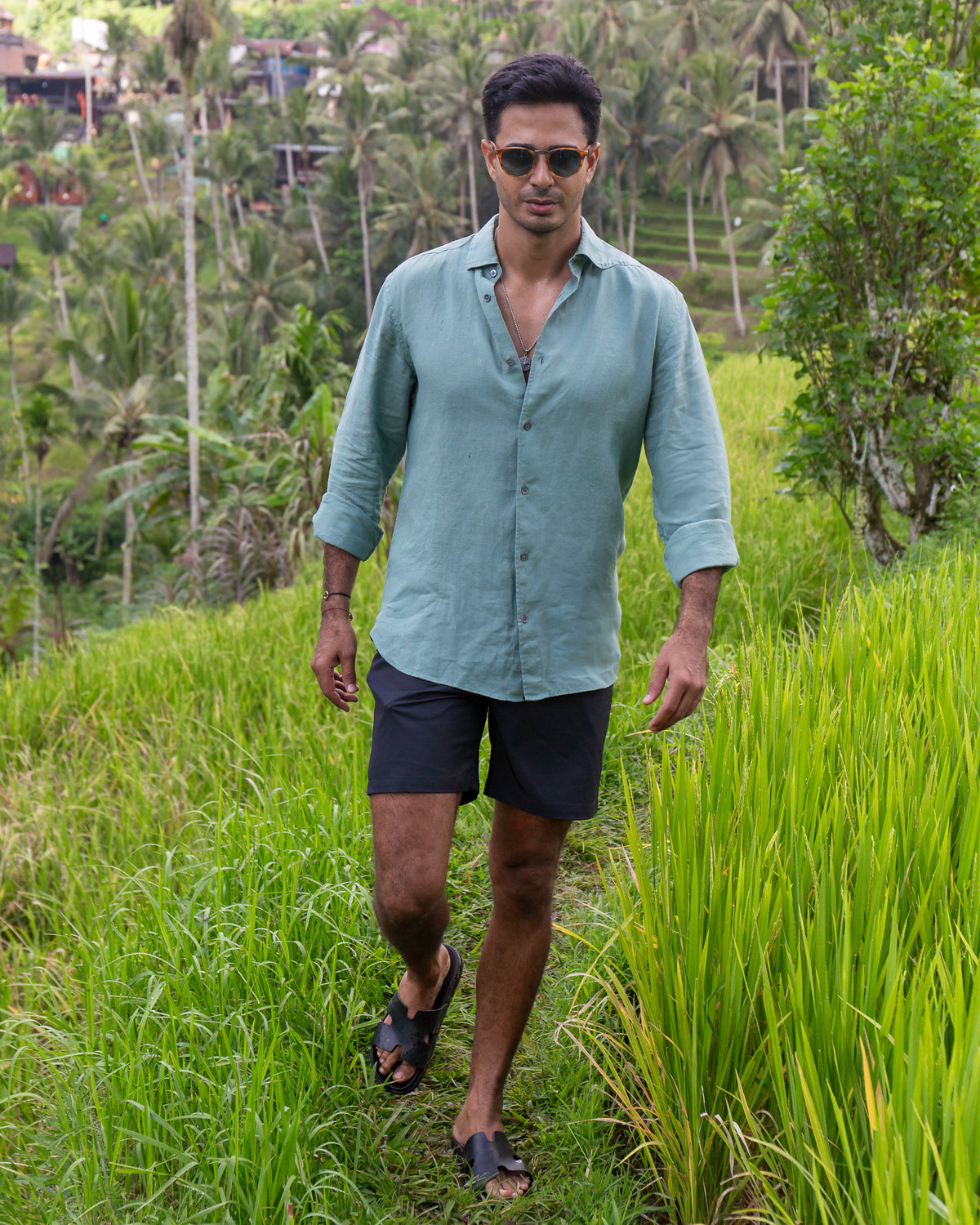 A man walking through a lush green rice field wearing a light RWENZORI sage green linen button-up shirt, dark shorts, and sunglasses.