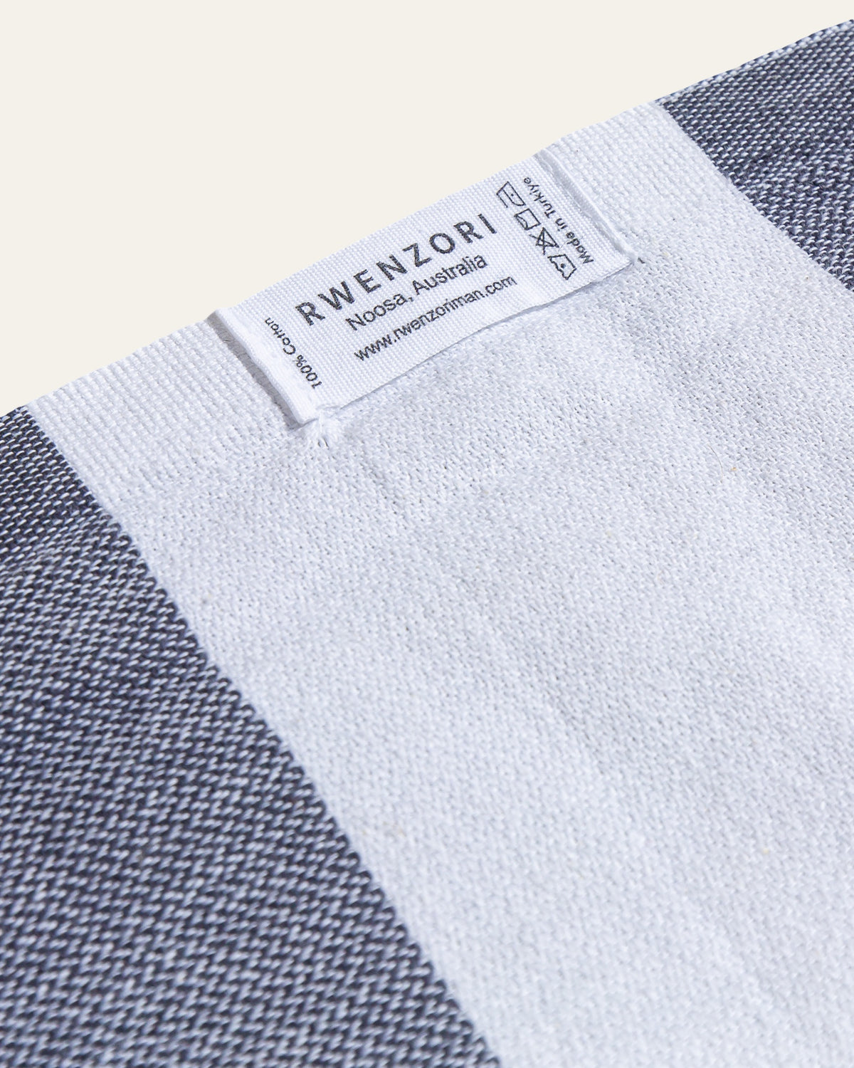Close-up of a woven textile product showing a white sewn-in label that reads RWENZORI, with a light grey woven pattern visible along the edges on a warm off-white background.