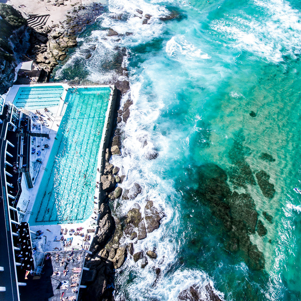 Aerial view of Bondi Beach Australia and outdoor pool with turquoise water, surrounded by rocky shoreline and crashing waves.