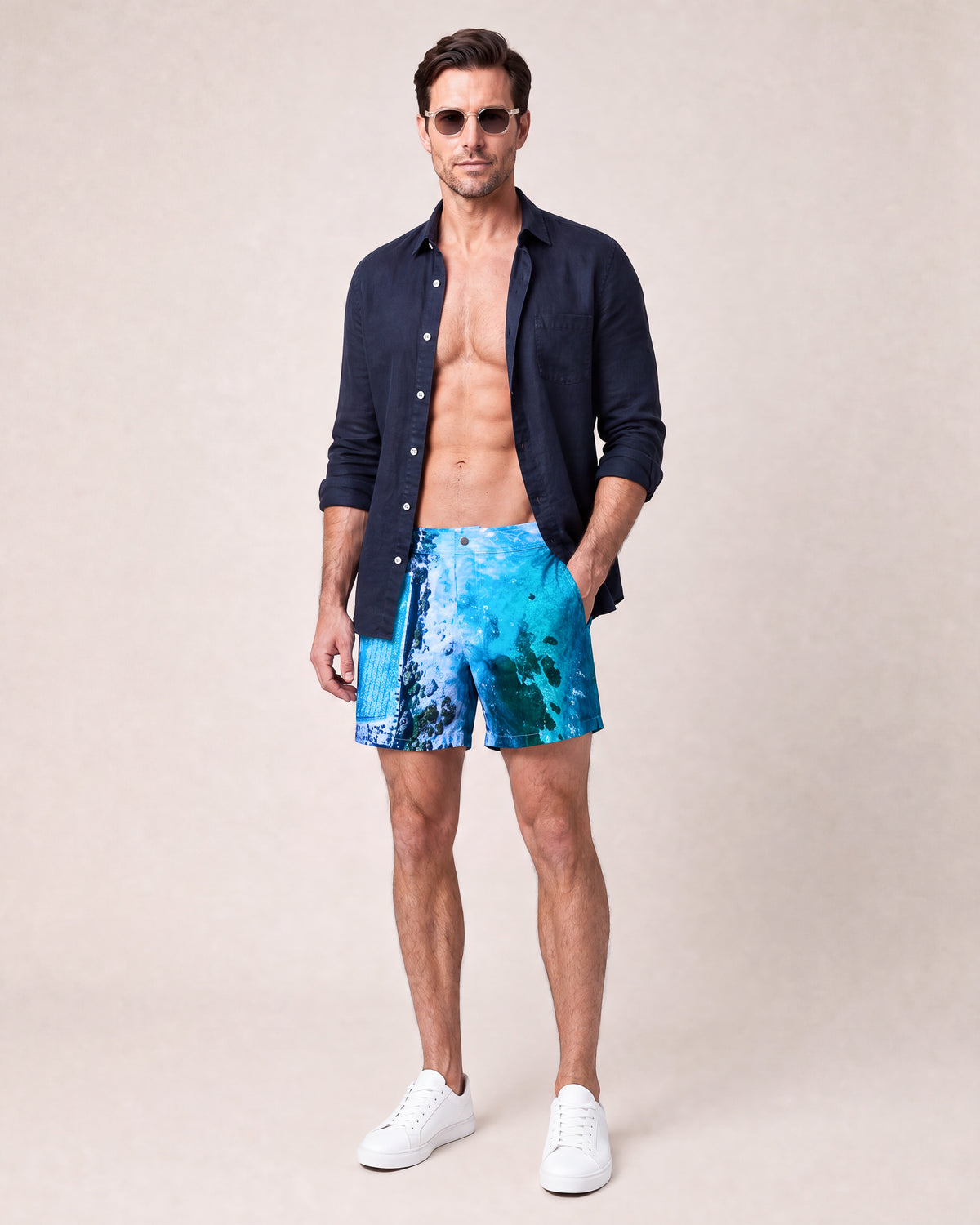 Man wearing RWENZORI Bondi photo print swim shorts with an open navy linen resort shirt.