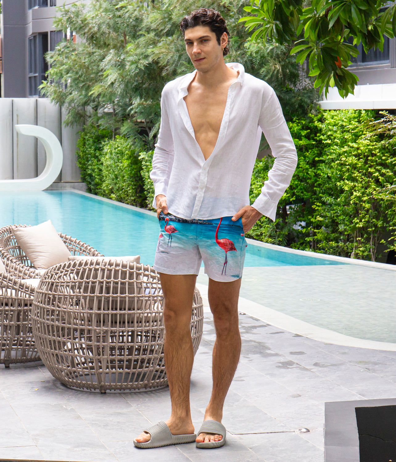 Man wearing a white linen shirt and Rwenzori flamingo-patterned swim shorts poolside.