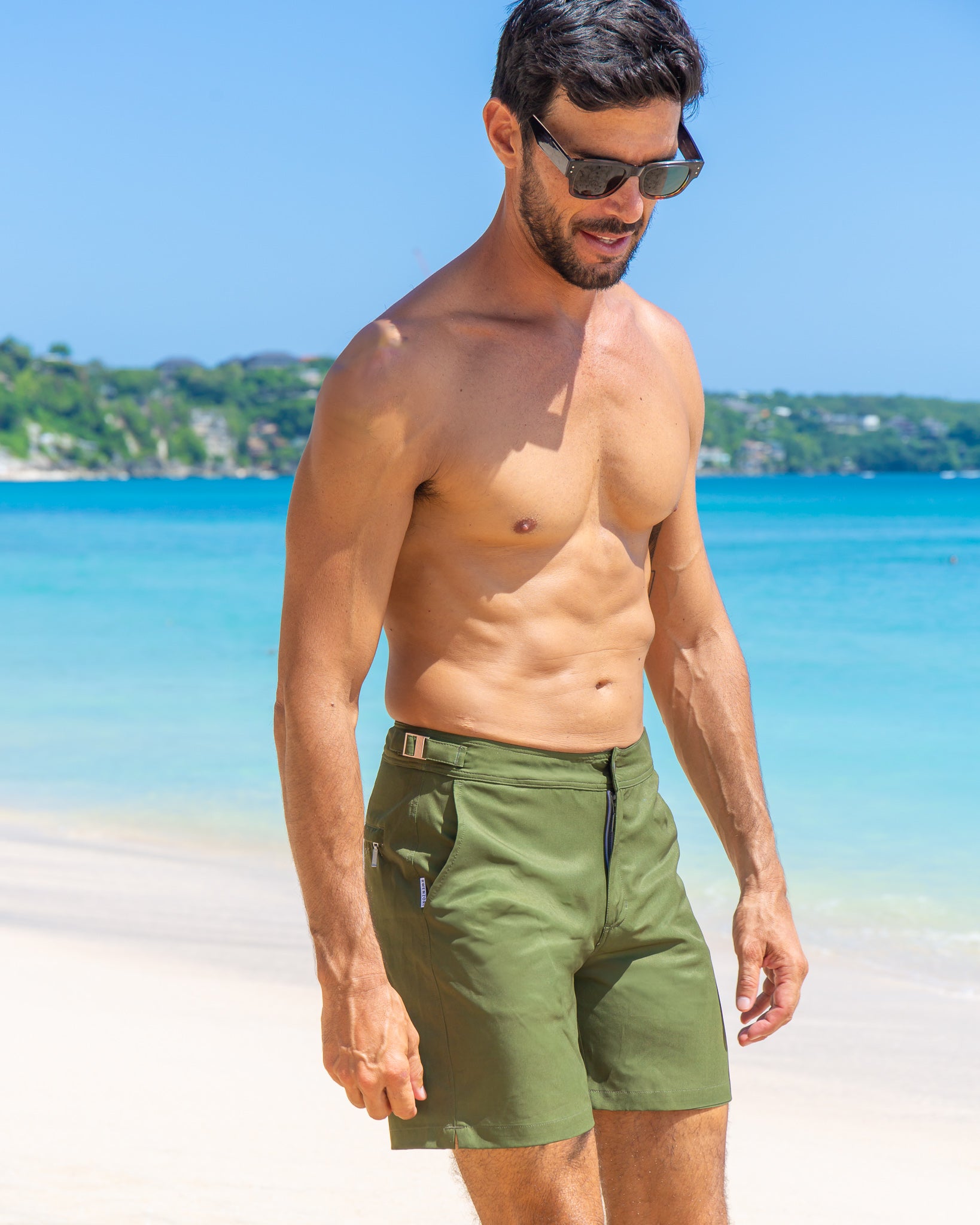 Man styled for the beach wearing RWENZORI Tuscan Olive Performance Series swim trunks with blue ocean water and white sand background.